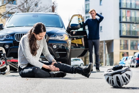 A dirt path running through a corn maze between tall stalks of green and golden corn, with a wooded hill of fall foliage and a bright blue sky in the background.