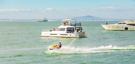 A dirt path running through a corn maze between tall stalks of green and golden corn, with a wooded hill of fall foliage and a bright blue sky in the background.